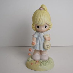 Precious Moments figurine of a young blond girl holding a bouquet of tiny hearts standing on a small patch of ceramic grass and flowers. Wearing a pretty blue dress and carrying a seed bag